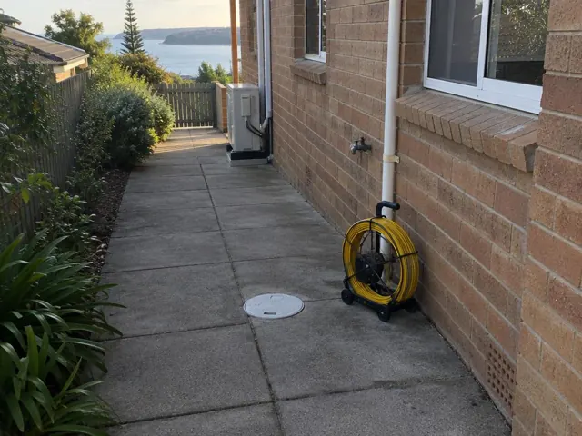 Capped inspection opening beside a yellow drain camera reel on a paved side path by a brick house in Cronulla
