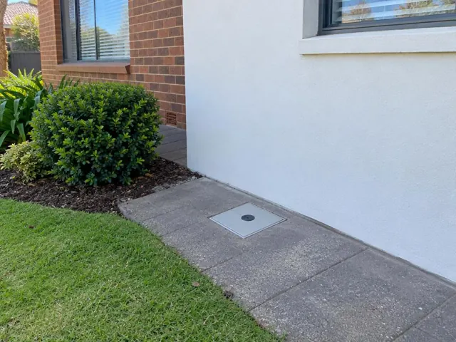 Flush capped access cover in concrete paving beside a brick and rendered house in Miranda